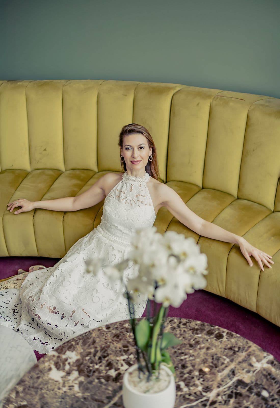Chen Reiss in a white lace dress, seated on a mustard velvet sofa